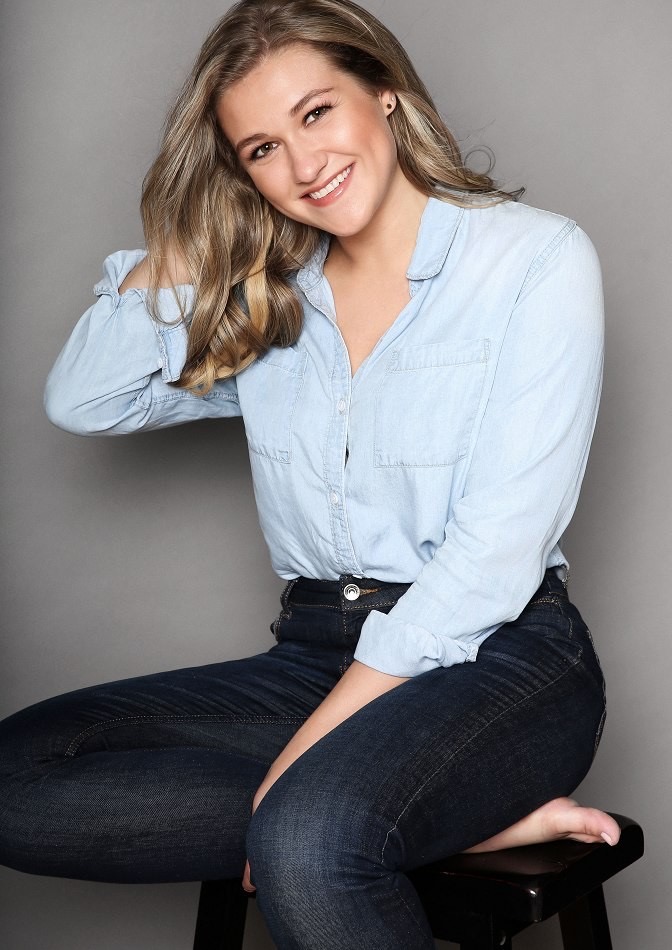 Kari Emerson smiling in a light blue denim shirt and dark jeans, seated on a stool